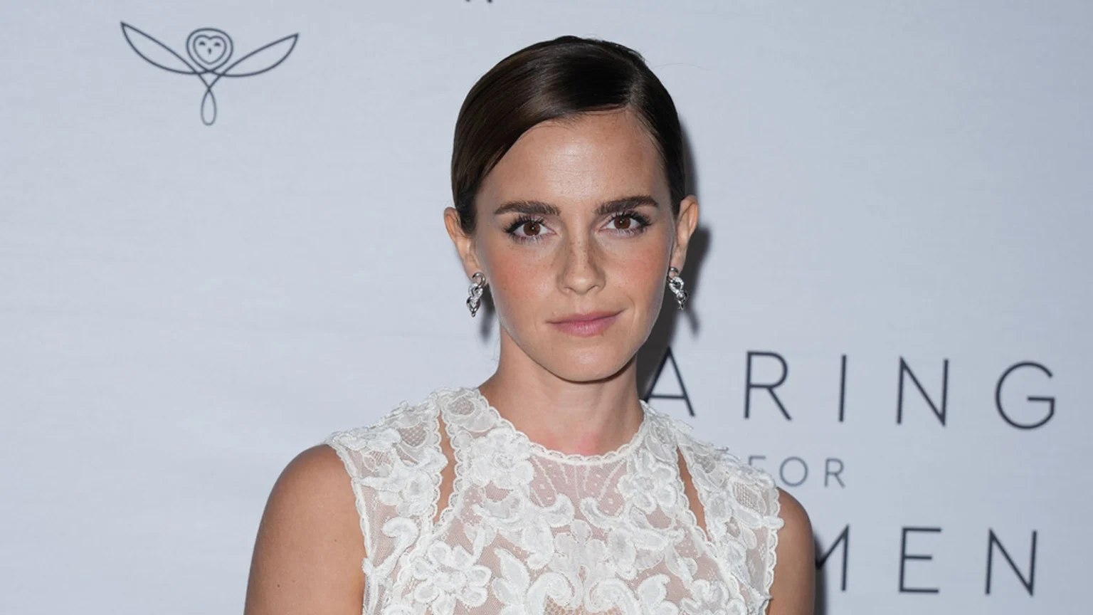 Emma Watson looks directly at the camera, with her hair neatly pulled back, wearing a white lace top and silver earrings. A stylized owl logo and partial text "ARING FOR MEN" are visible in the light background.