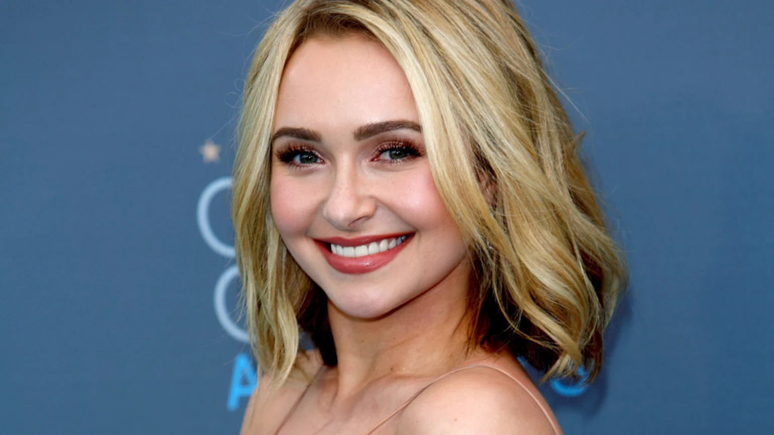 Close-up of actress Hayden Panettiere smiling, with blonde wavy hair, green eyes, and light makeup, against a soft blue background with faint award ceremony branding.