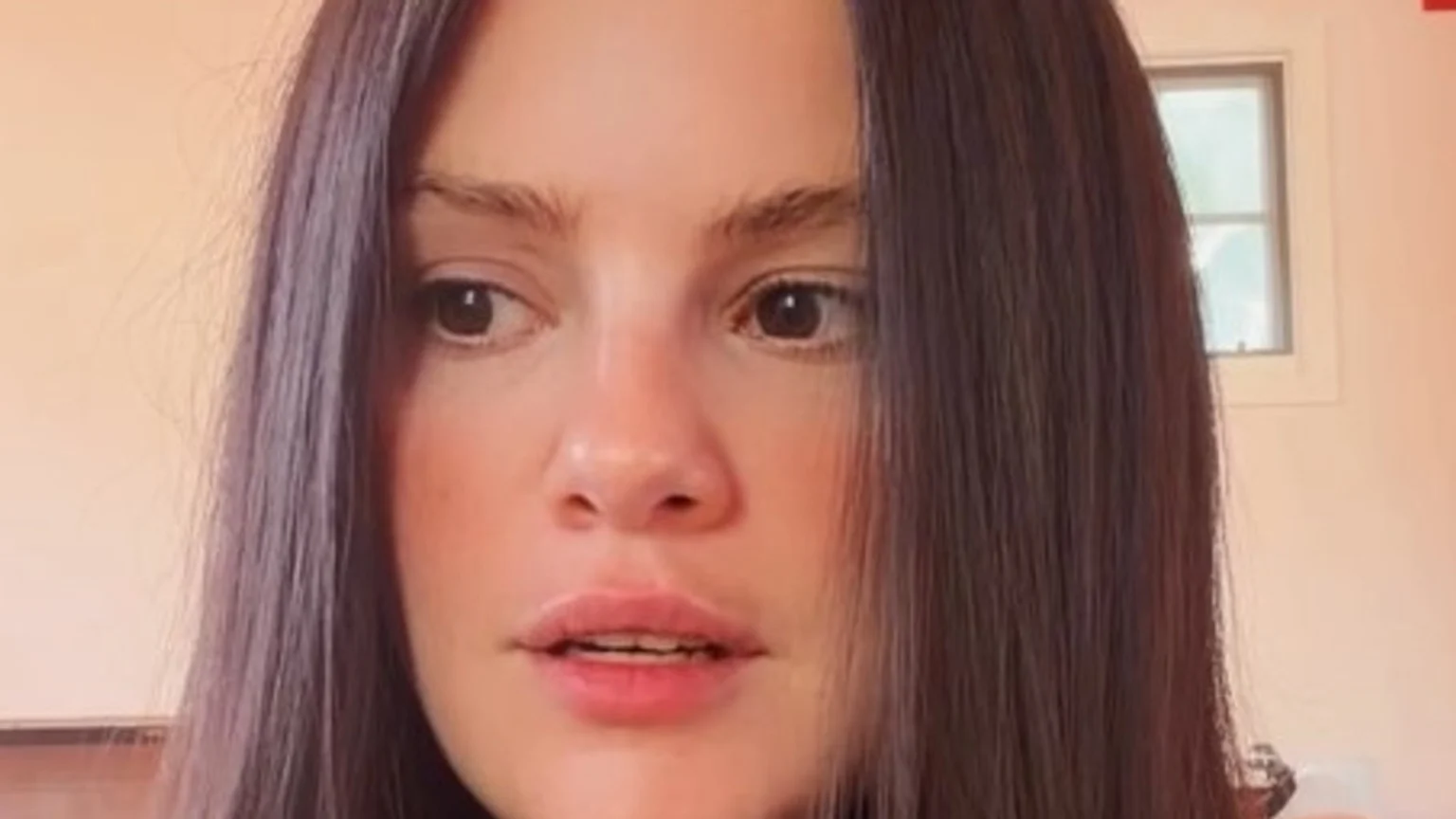 A close-up of a woman with long dark hair, her eyes looking to the side and her lips slightly parted showing her bottom teeth, against a light wall with a window.