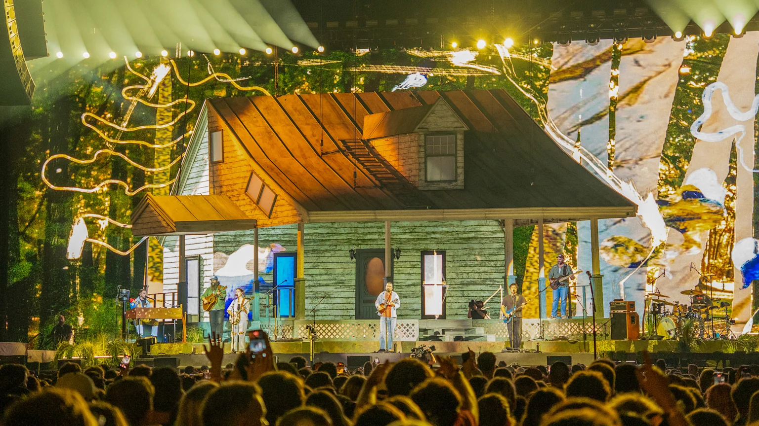 Musicians perform on a stage designed as a rustic house with a porch, against a large video screen displaying forest scenes, illuminated by spotlights, with a large crowd in the foreground.