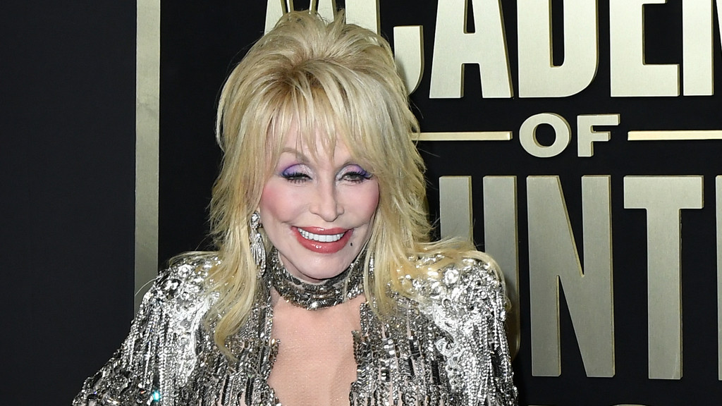 Dolly Parton smiles, wearing purple eyeshadow, pink lipstick, and a shimmering silver outfit with a choker and earrings, against a black and gold background.