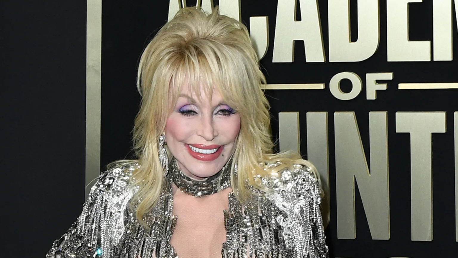 Dolly Parton smiles, wearing purple eyeshadow, pink lipstick, and a shimmering silver outfit with a choker and earrings, against a black and gold background.