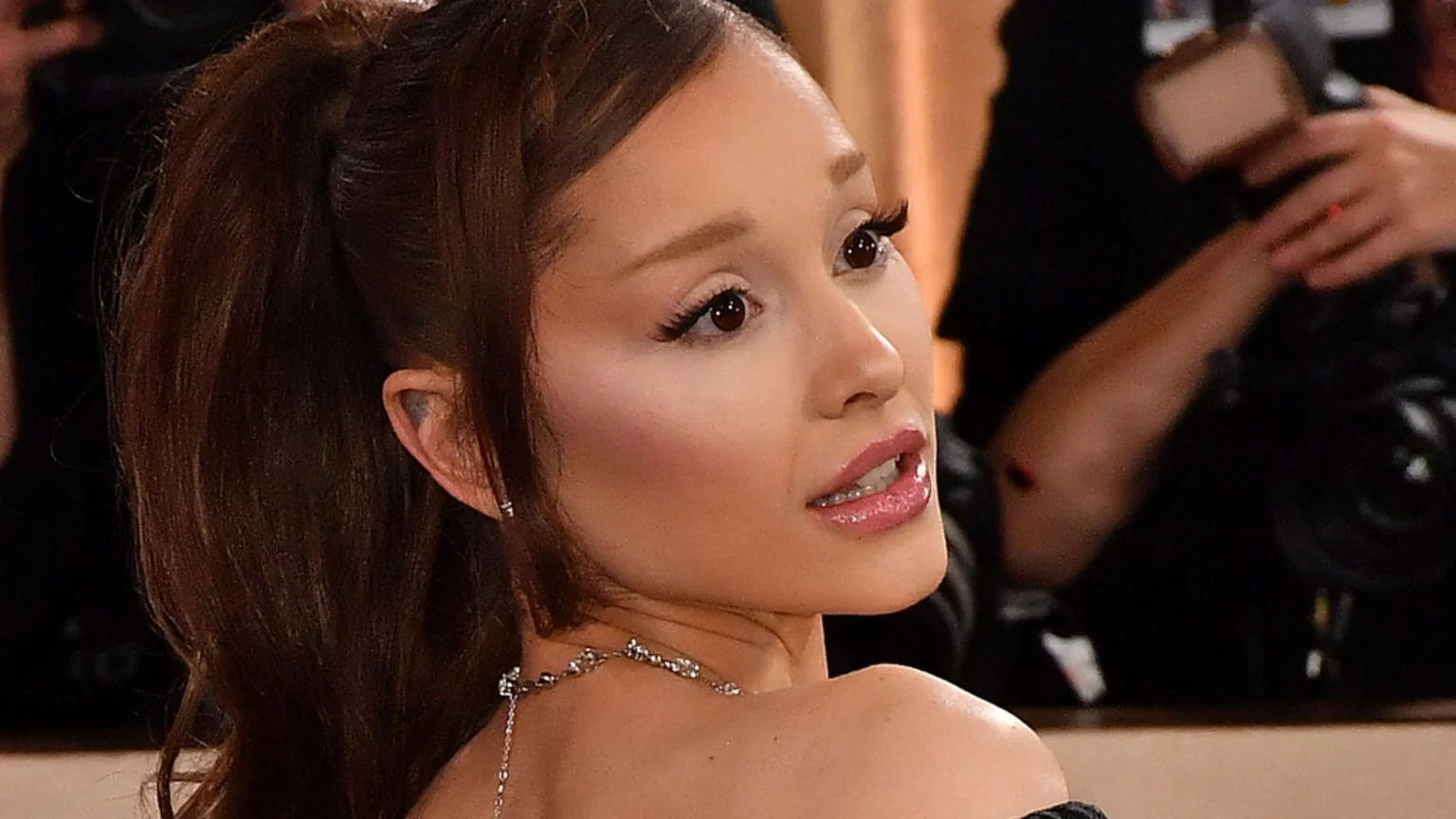 Ariana Grande looks over her right shoulder, with dark hair in a high ponytail, winged eyeliner, glossy pink lips, and a silver necklace.