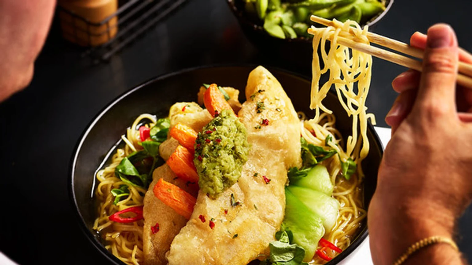 A person uses chopsticks to lift noodles from a black bowl of ramen containing fried fish, green sauce, vegetables, chili, and orange pieces, with a bowl of edamame in the background.