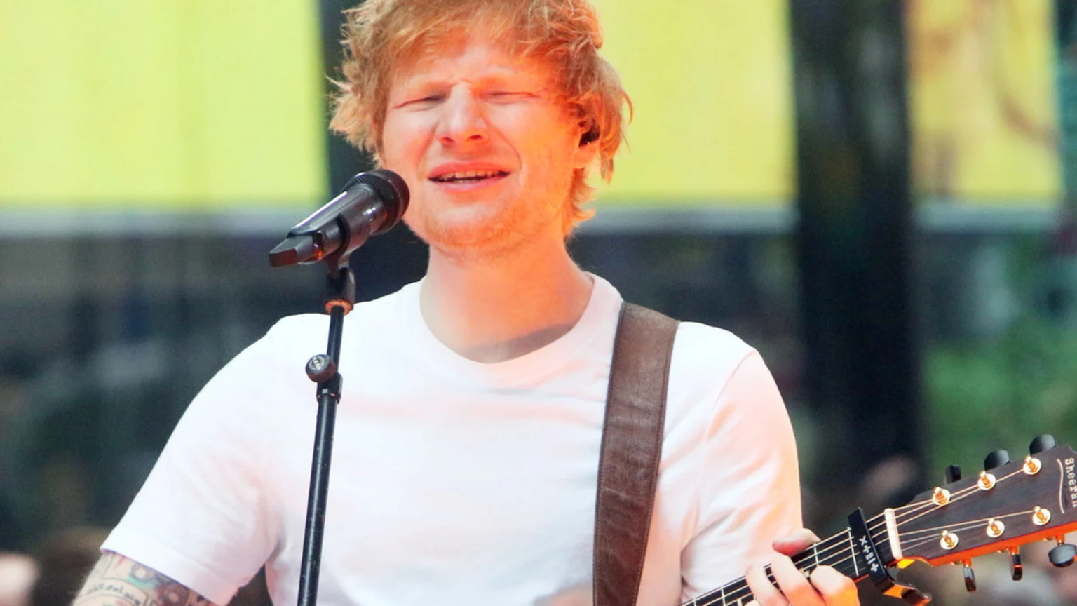 Ed Sheeran, with red hair and tattoos on his left arm, sings passionately into a microphone with his eyes closed, holding an acoustic guitar, and wearing a white t-shirt against a blurred background.