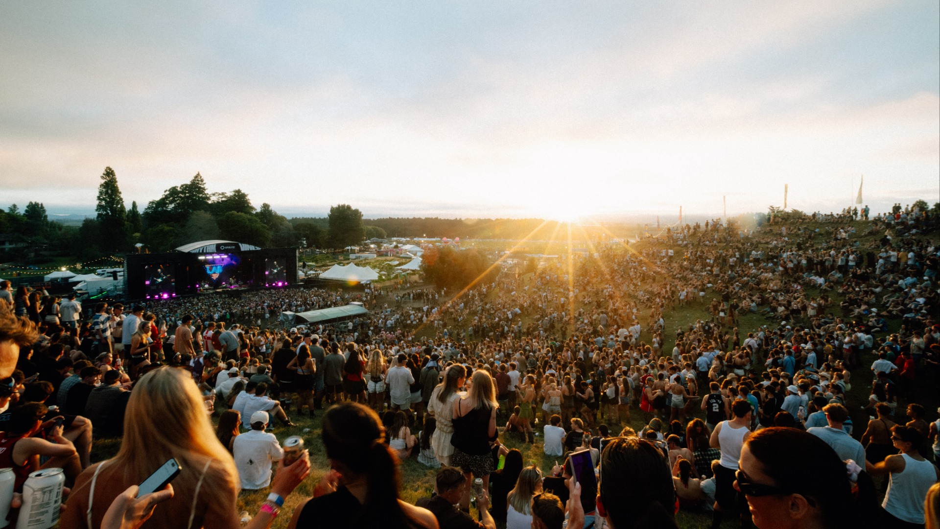 A lively outdoor music festival scene at sunset, showing a large crowd of people on a grassy hill watching a brightly lit stage, with the sun's golden rays shining across the landscape.