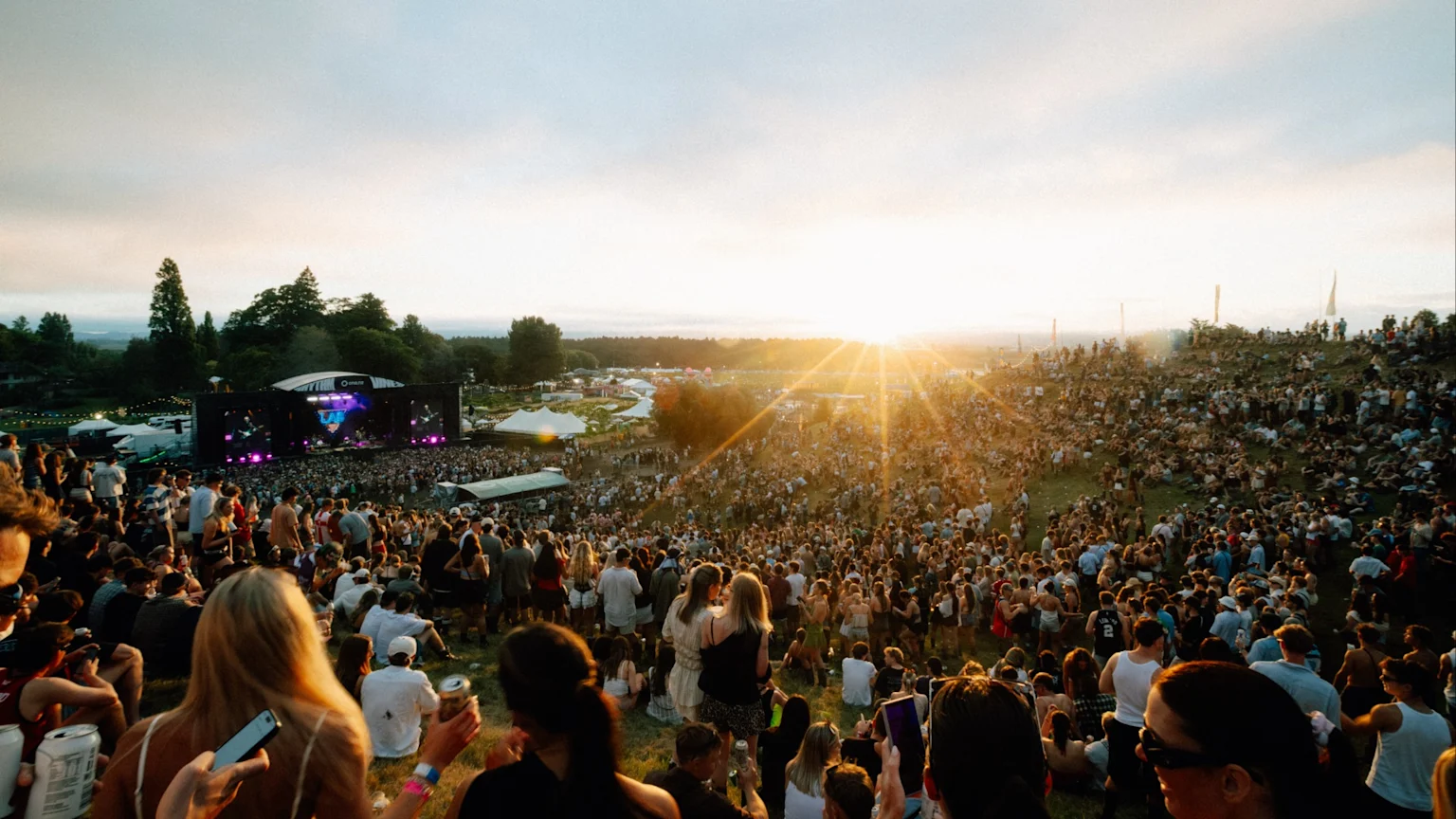 A lively outdoor music festival scene at sunset, showing a large crowd of people on a grassy hill watching a brightly lit stage, with the sun's golden rays shining across the landscape.