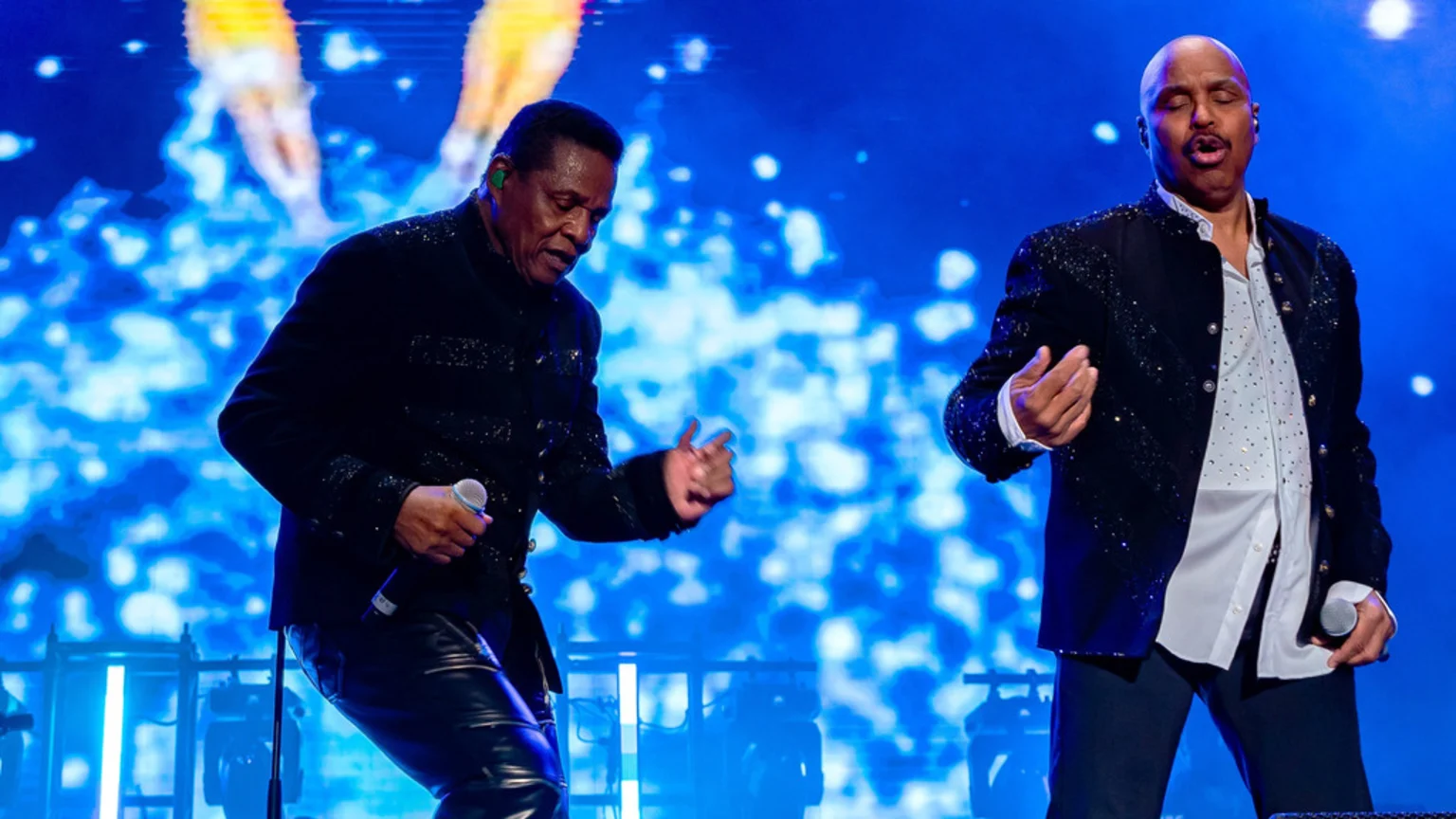 Two male performers, dressed in sparkling dark jackets, sing into microphones on a stage with a vivid blue background featuring abstract light patterns and glowing stage equipment.