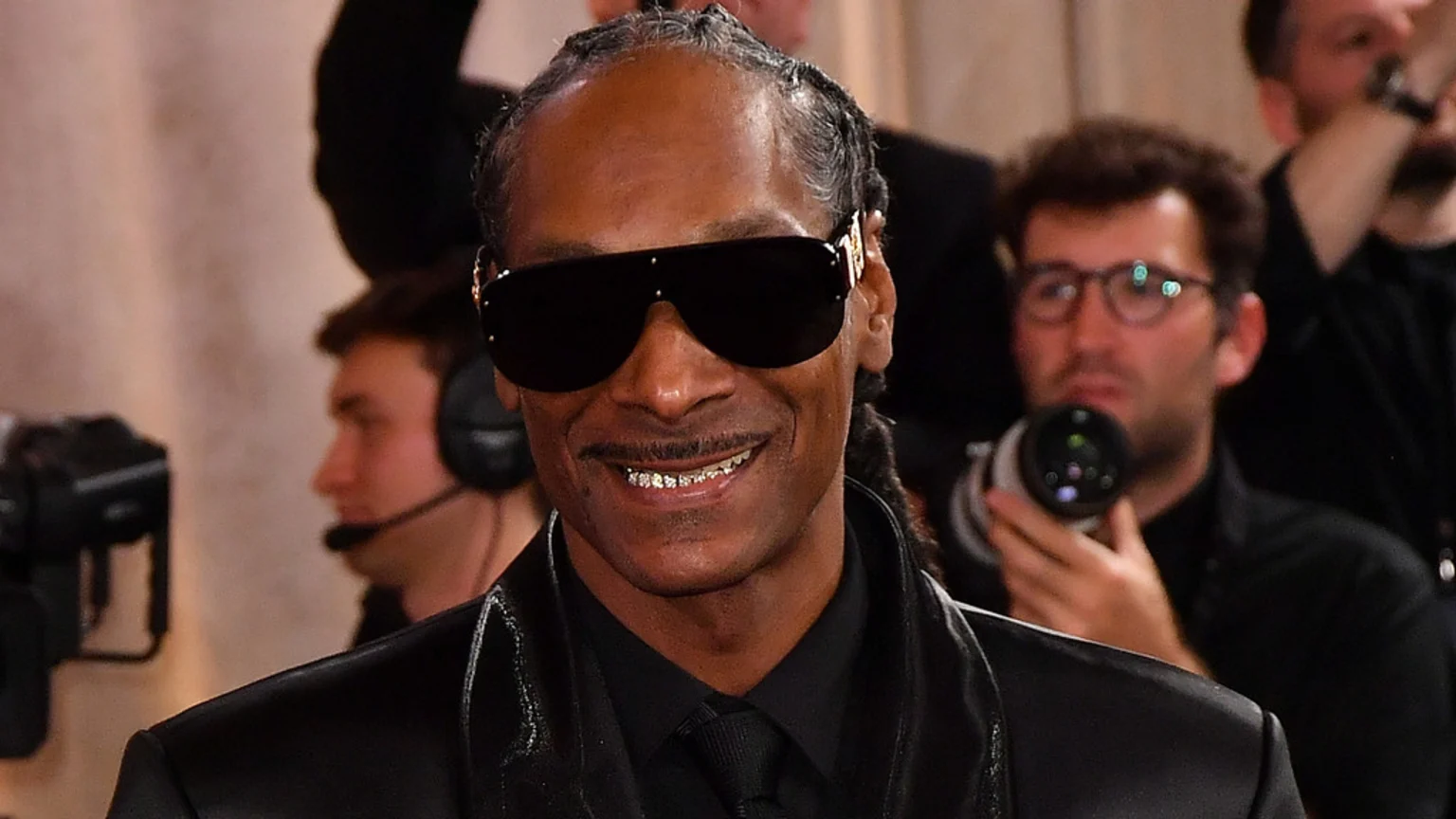 Snoop Dogg smiles, wearing dark sunglasses, a black suit, and showing grills on his teeth, with blurred people and cameras in the background.