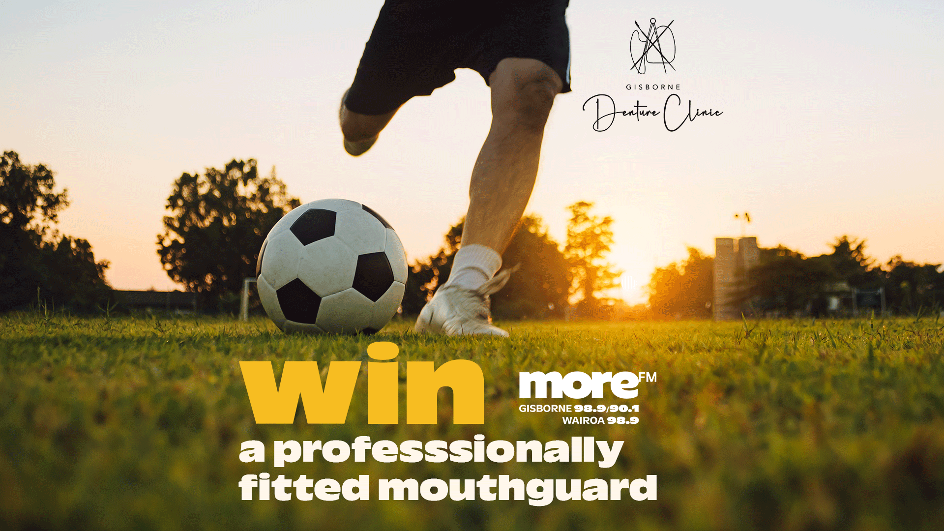 A person's legs are poised to kick a soccer ball on a grassy field at sunset, with text "WIN a professsionally fitted mouthguard" promoted by "Gisborne Denture Clinic" and "more FM".