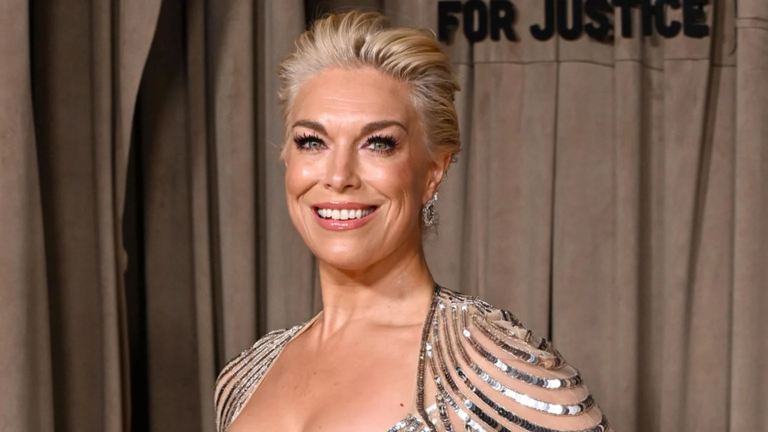 Hannah Waddingham smiles brightly, looking at the camera, with blonde hair styled up and wearing a sparkly, sequined top against a blurred brown curtain background that reads "FOR JUSTICE".