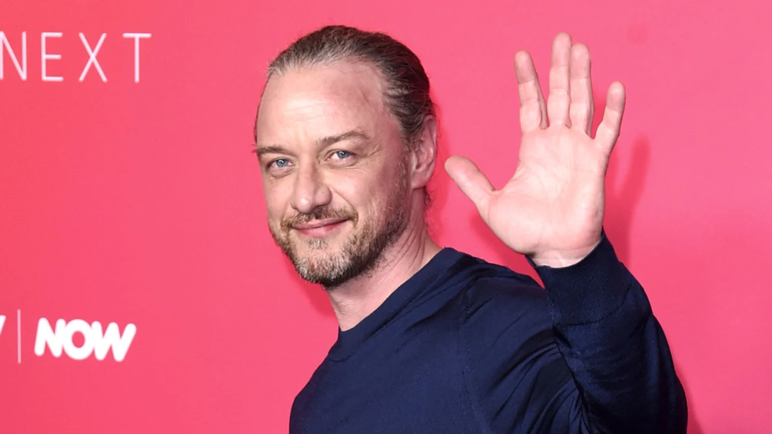 James McAvoy, with pulled-back hair and a beard, smiles and waves while wearing a navy blue shirt against a pink background with "NEXT" and "NOW" text.