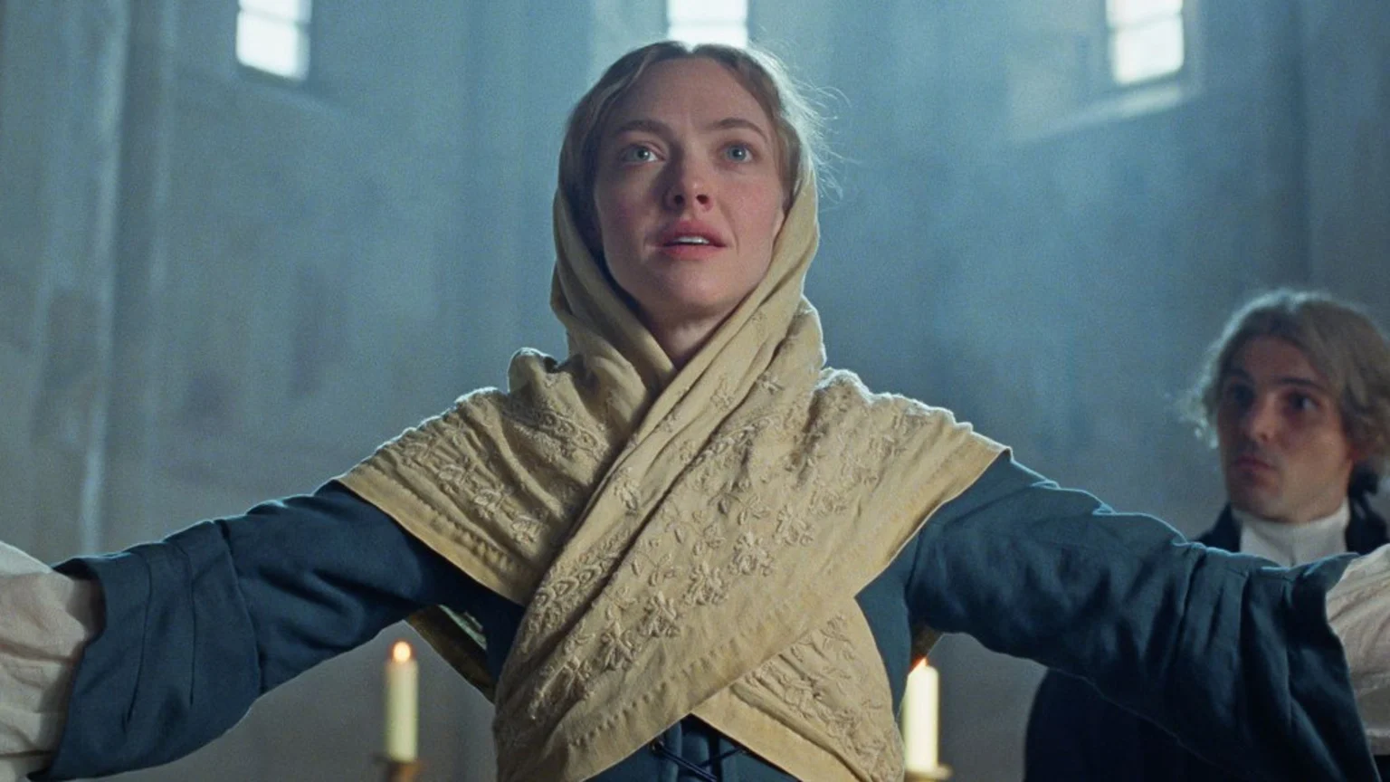 Amanda Seyfried, wearing a beige head covering and a blue dress, stands with arms outstretched and an earnest expression, looking upwards in a dimly lit room with candles and a man partially visible on the right.