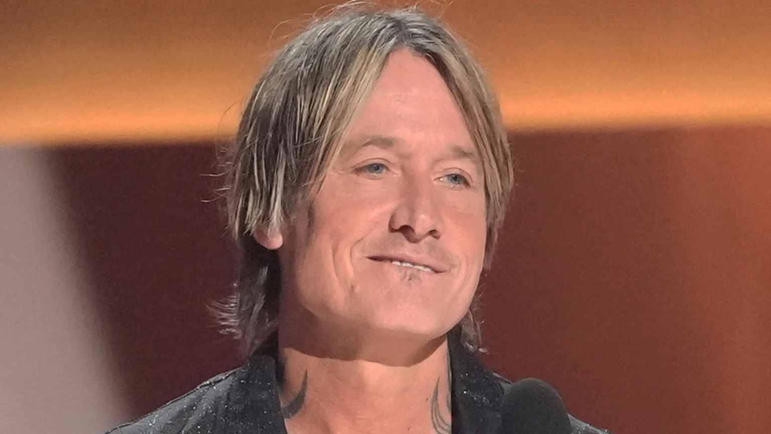Keith Urban with shoulder-length blond hair and blue eyes, smiling slightly, with neck tattoos visible, speaks into a microphone against a warm, gradient background.