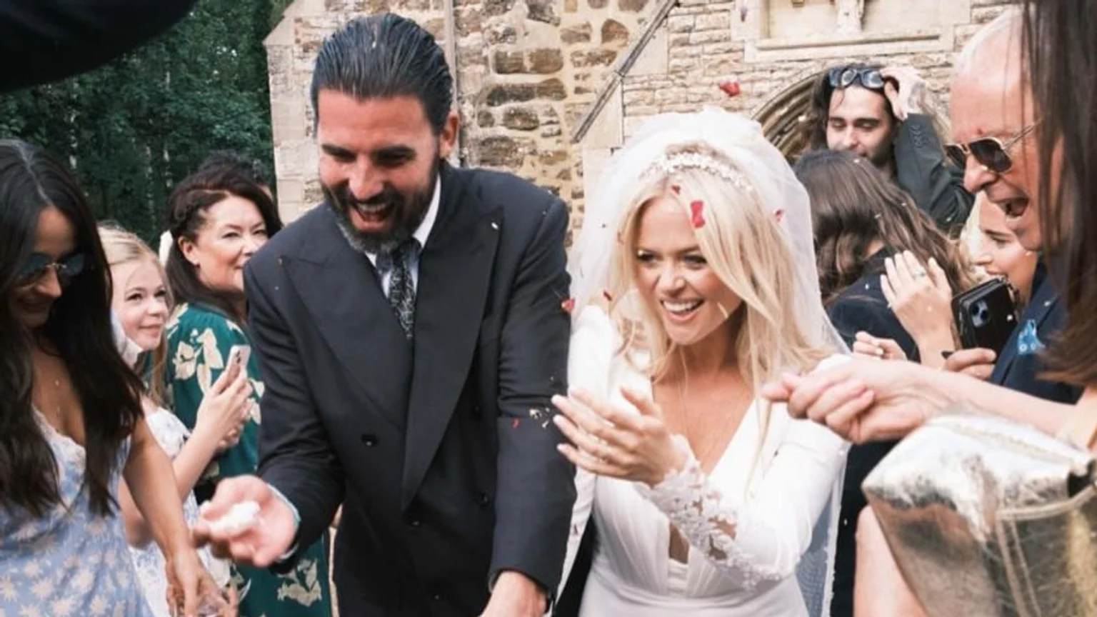 A smiling bride in a white dress and veil and a laughing groom in a dark suit are showered with confetti by cheerful guests outside a stone building.