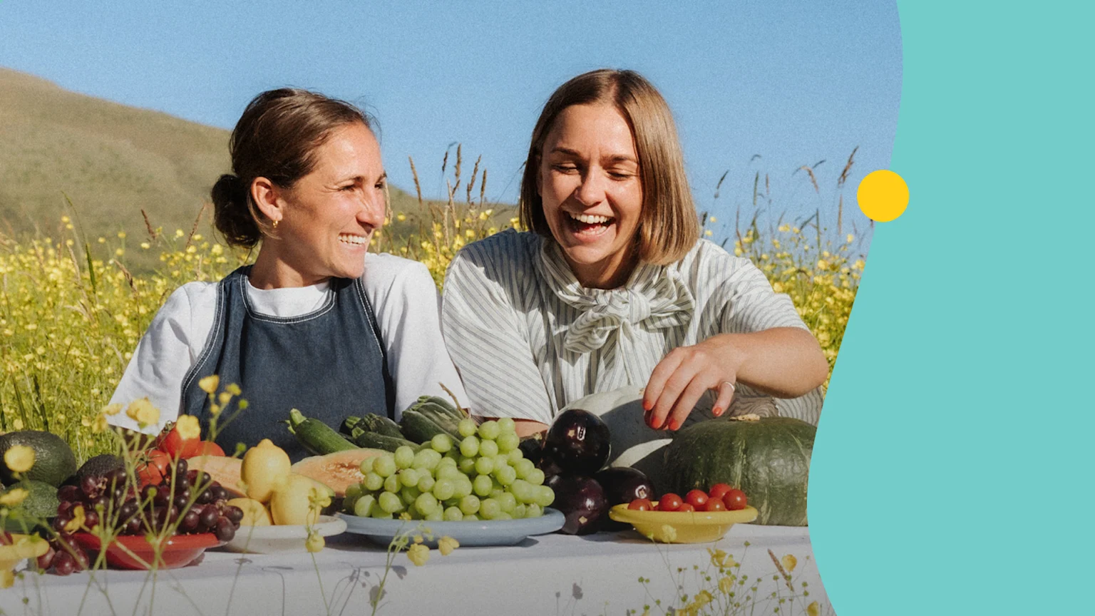 Two women, laughing and smiling, sit at a table laden with various fresh fruits and vegetables in a sunny, flower-filled field.