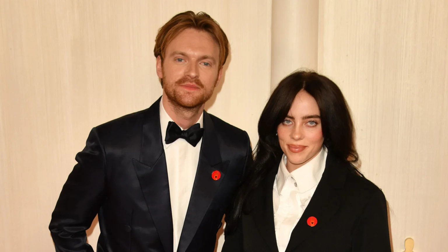Billie Eilish and Finneas O'Connell stand side-by-side, both wearing red pins on their dark formal attire, with Billie in a black blazer and Finneas in a dark suit and bow tie.
