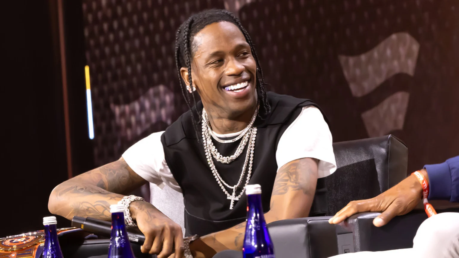 Travis Scott smiles, wearing braids, chains, and a black vest, while seated at a table with water bottles and a championship belt.