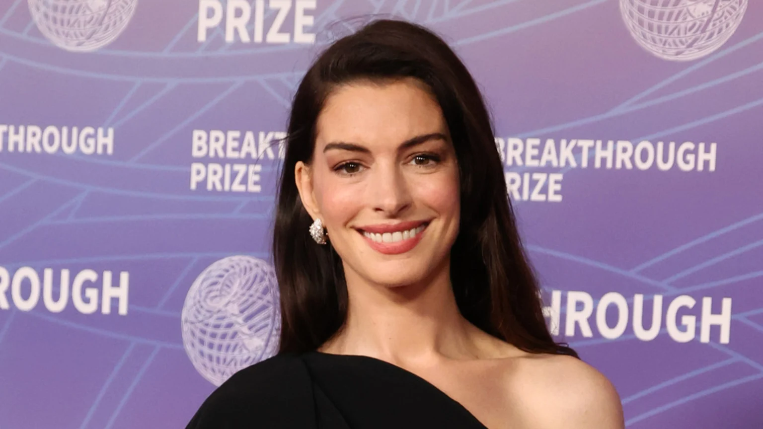 Anne Hathaway, with long dark hair, smiles while wearing a black one-shoulder dress and diamond earrings against a purple background with "Breakthrough Prize" text.