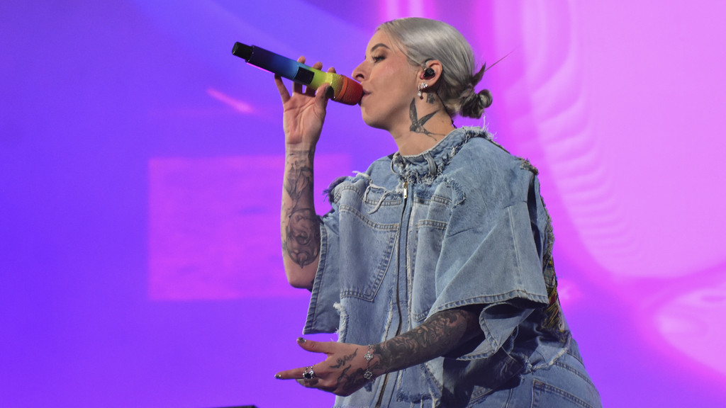 A person with light gray hair and arm tattoos sings into a rainbow microphone, wearing a denim top against a purple background.