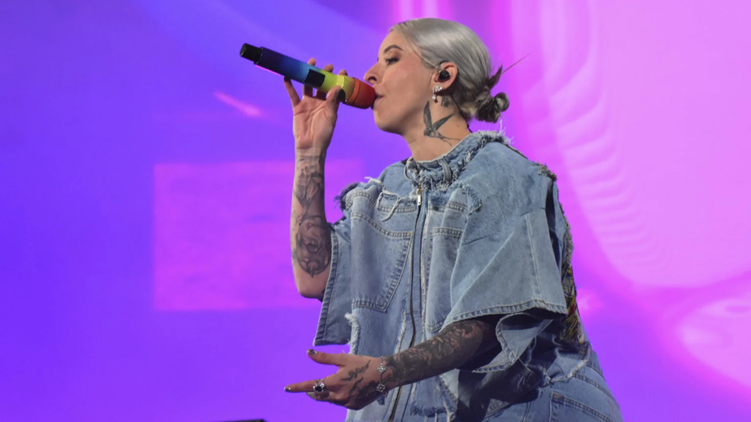 A person with light gray hair and arm tattoos sings into a rainbow microphone, wearing a denim top against a purple background.