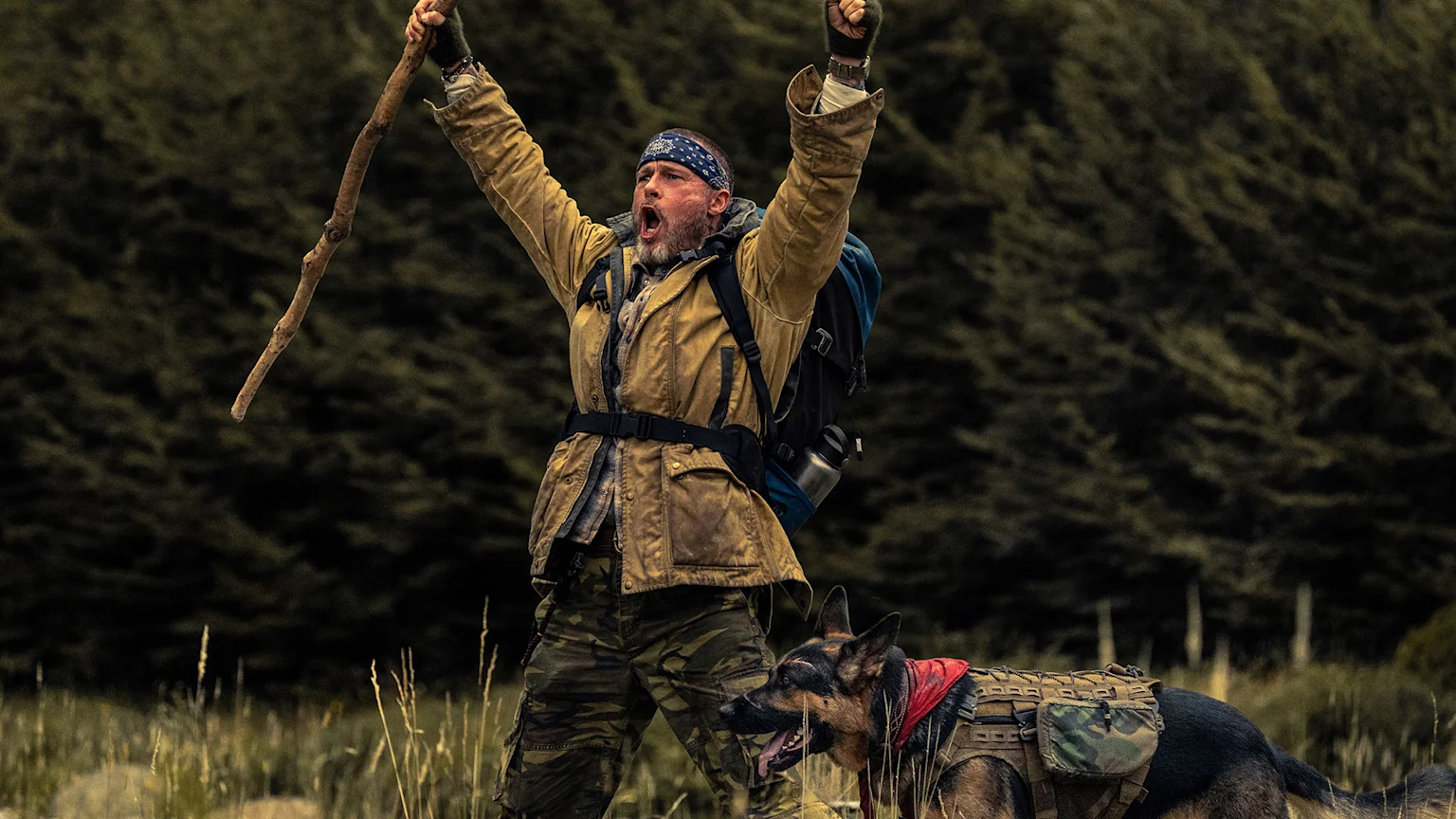 A man in outdoor gear with a backpack and stick raises his arms triumphantly and shouts, accompanied by a German Shepherd dog wearing a tactical vest, in a forest.