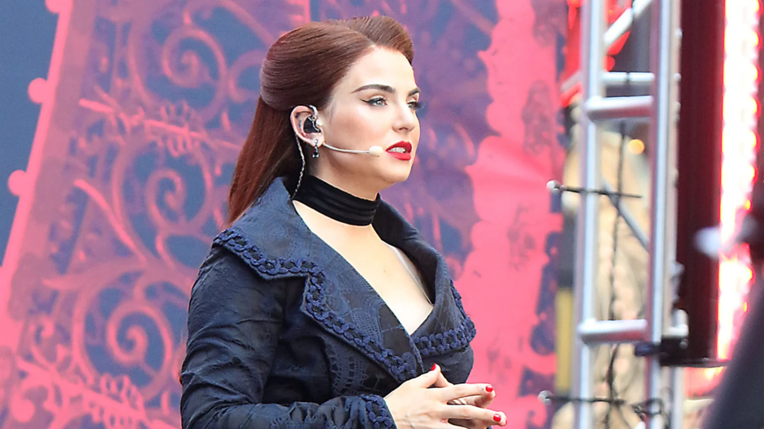 A woman with reddish-brown hair, red lipstick, and a dark ornate jacket with blue trim wears an in-ear monitor and microphone, looking focused on a stage with a blurry red background.