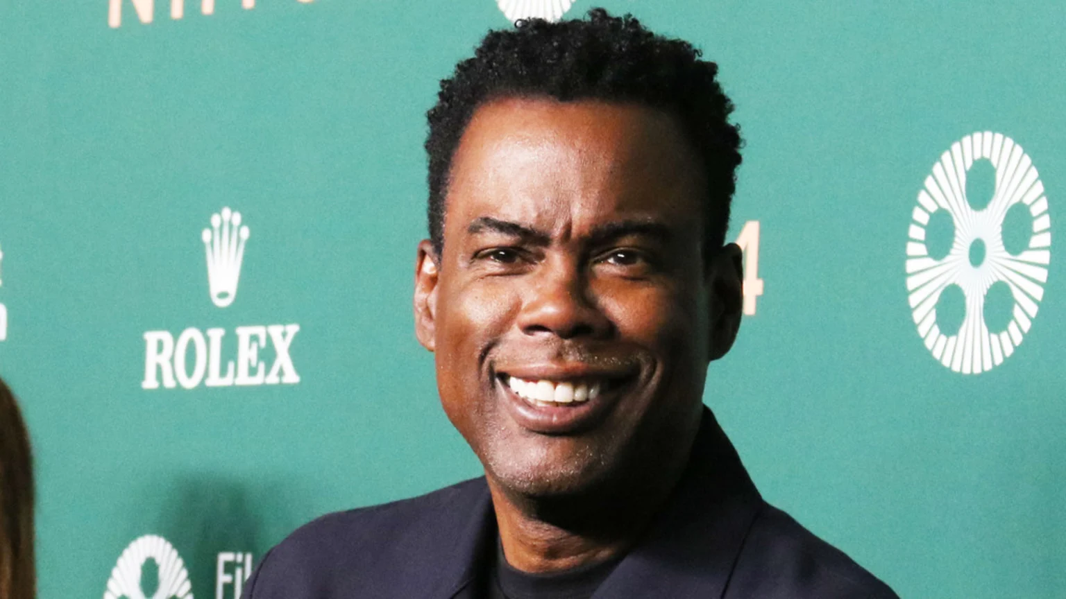 Chris Rock, a Black man with short curly hair, smiles broadly, wearing a dark jacket against a teal backdrop featuring white Rolex and film reel logos.