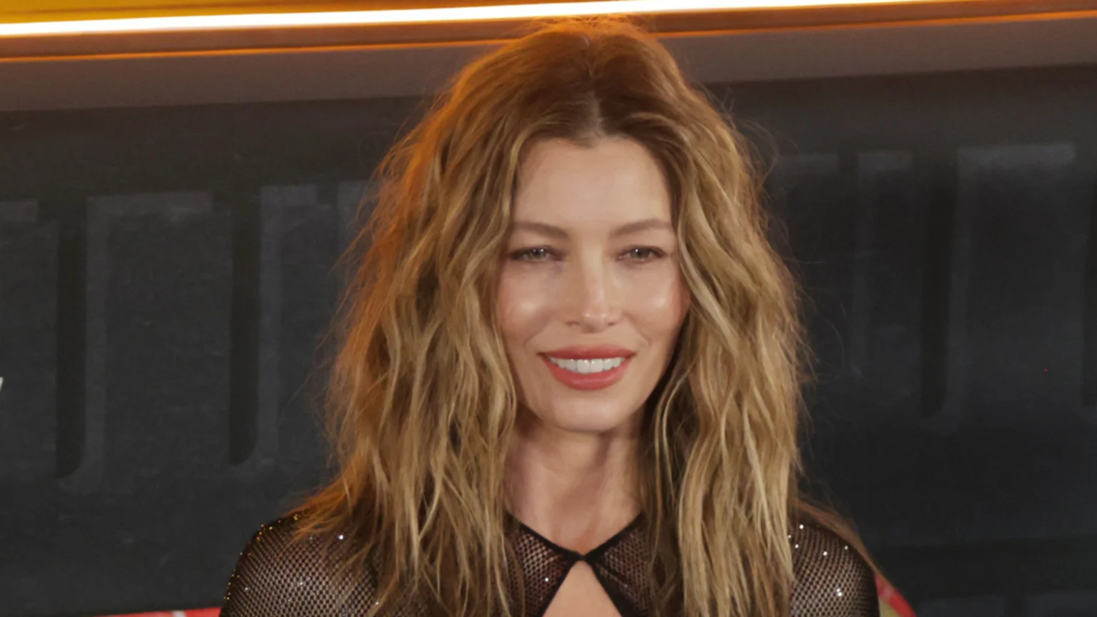 Jessica Biel smiles at the camera, wearing a black mesh top with a cutout and glitter, and long wavy blonde hair, against a dark background with orange lighting.