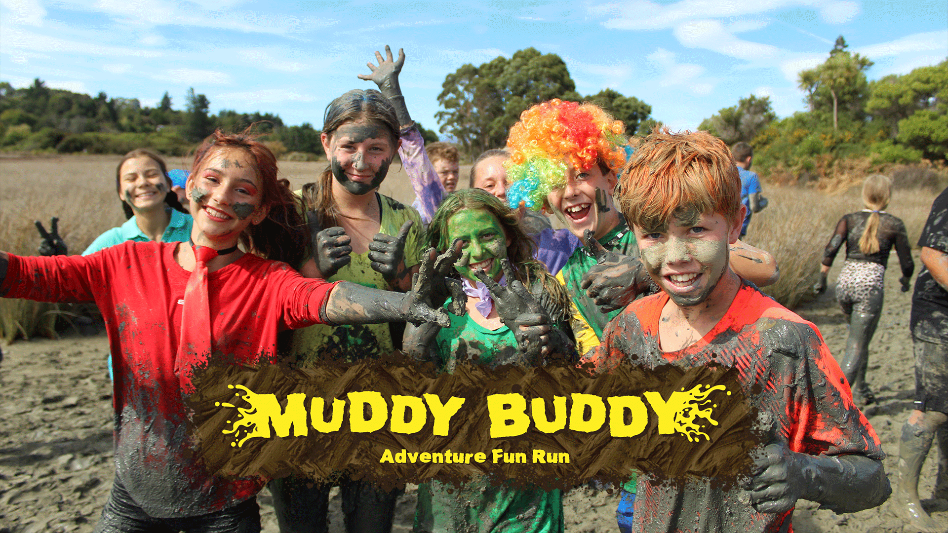 A group of excited kids, covered in mud, smile widely while participating in the "Muddy Buddy Adventure Fun Run" in an outdoor, muddy setting.