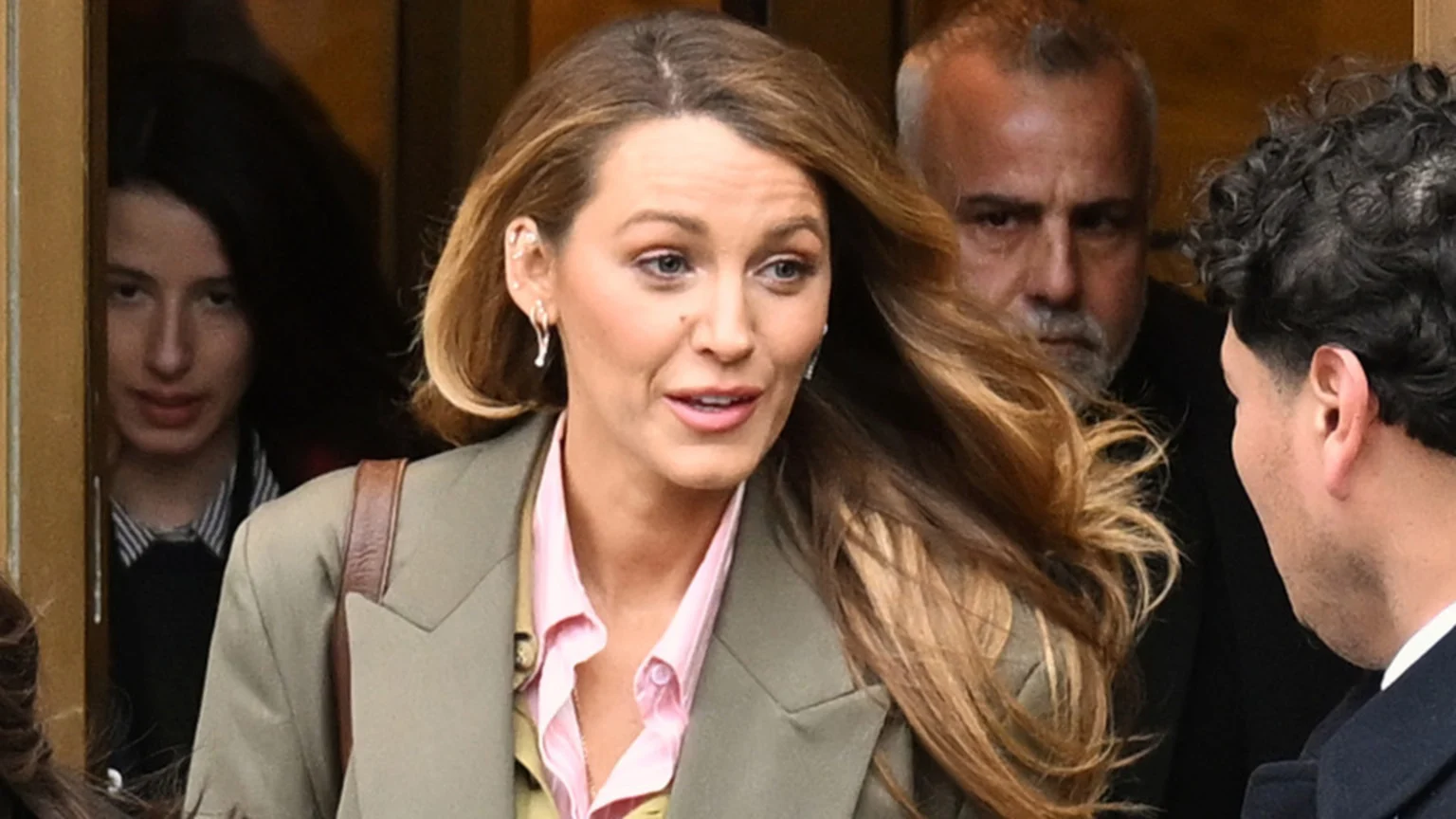 Blake Lively, with long wavy hair, wears a pink shirt and green blazer, looking and speaking to her left. Behind her, a grey-bearded man looks intently, a young person peeks from a dark doorway, and the back of another man's head is visible.