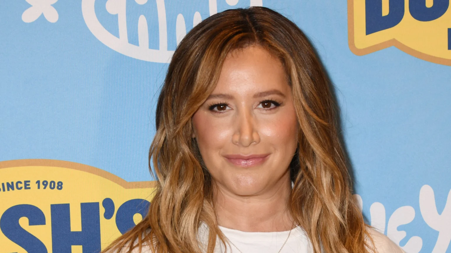 Ashley Tisdale, with long wavy brown hair, smiles subtly while looking at the camera against a light blue background with partially visible yellow and white logos.