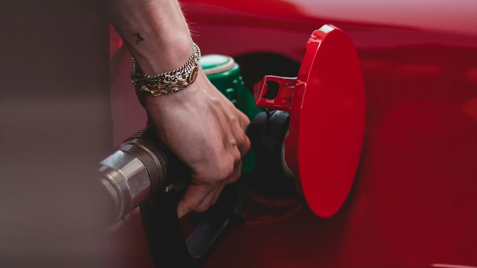 A hand with bracelets and a small tattoo inserts a green and black fuel nozzle into the open fuel tank of a red car, with the red fuel door visible.