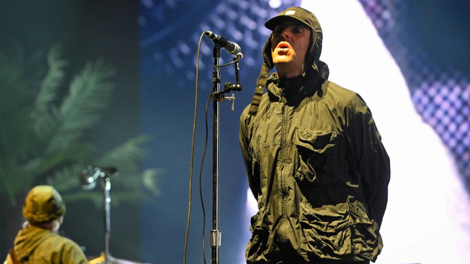 A man in a dark hooded jacket and baseball cap sings into a microphone on stage, with blurred stage lights and green shapes in the background.
