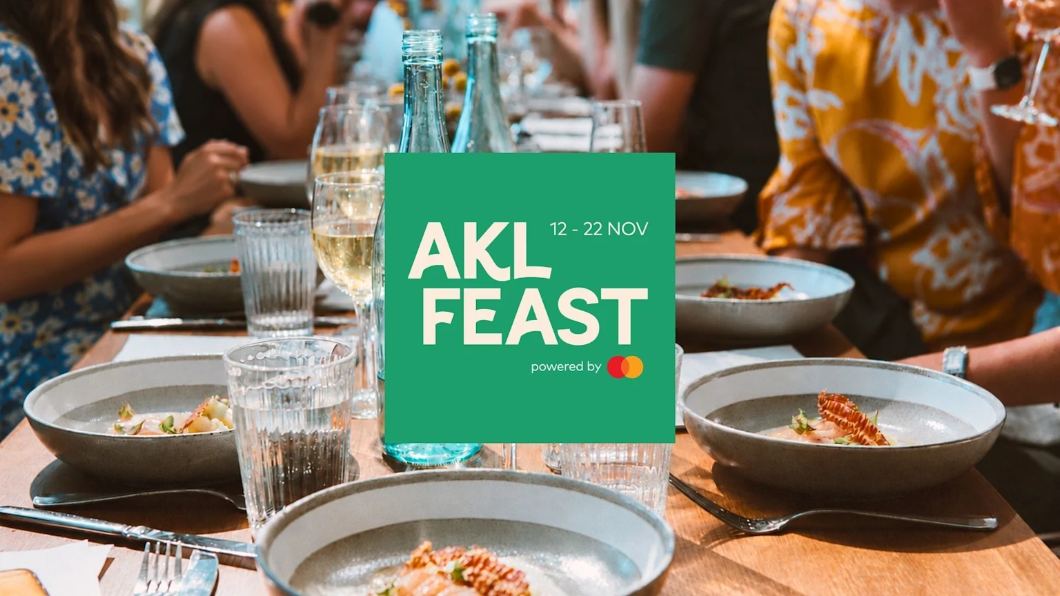People are dining at a long table with plates of elegant food and drinks, with a prominent green overlay displaying "AKL FEAST 12 - 22 NOV powered by Mastercard".