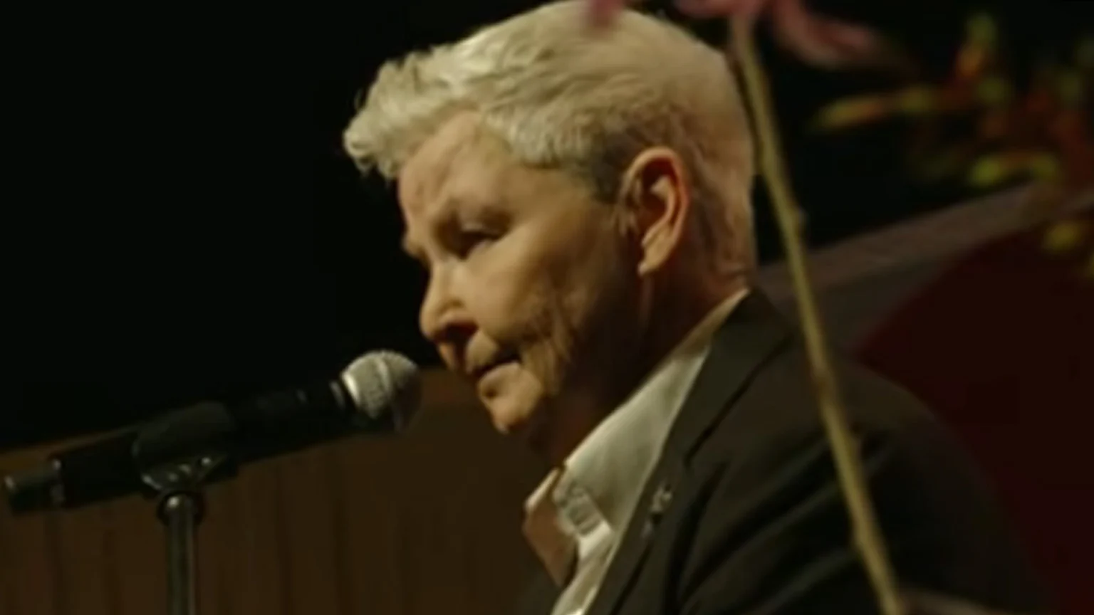 An older person with short, silver hair wears a dark jacket and white shirt, looking down at a microphone in profile against a dark, blurred background.