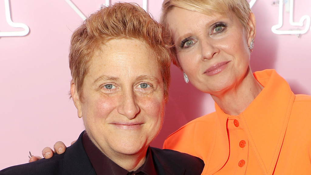 Cynthia Nixon, with short blonde hair and an orange top, smiles alongside a person with short reddish hair in a dark suit, her hand resting on their shoulder.