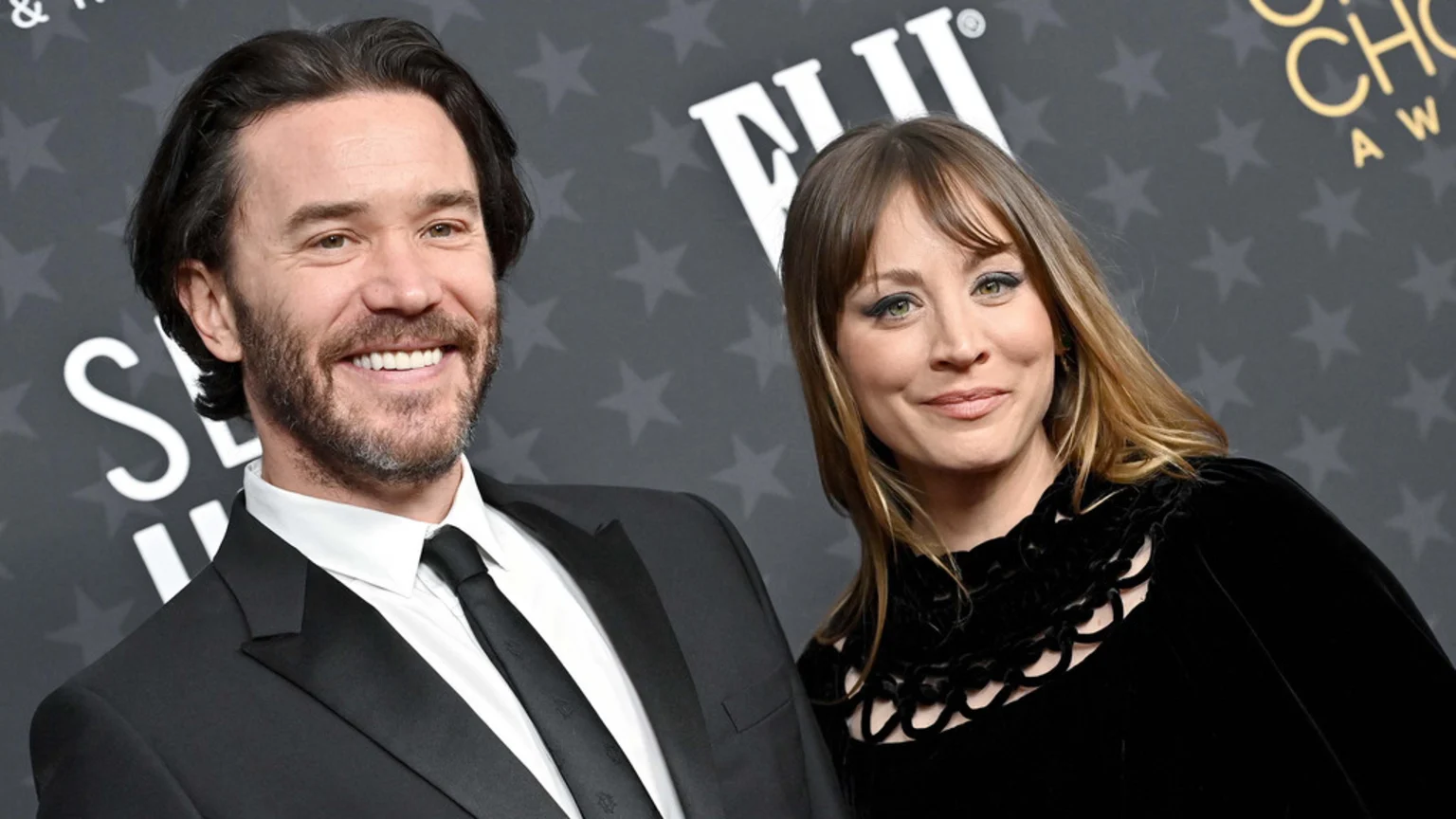 Kaley Cuoco, wearing a black dress, and Tom Pelphrey, in a black suit, smile at an event with a star-patterned background.