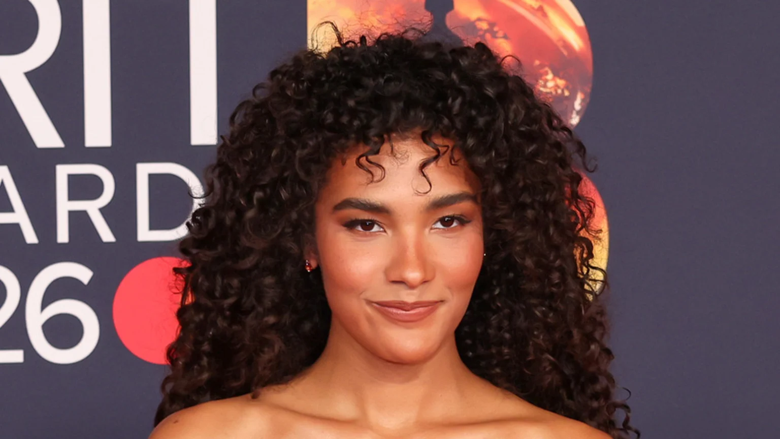 A woman with dark curly hair and light makeup smiles, with a blurred background showing parts of a "BRIT AWARDS" sign and orange circular graphics.