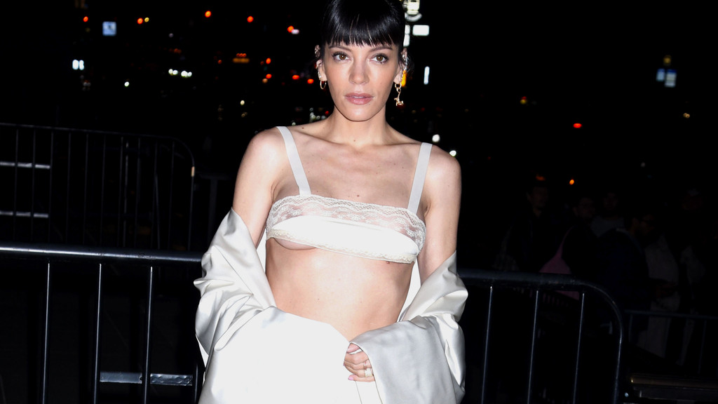 Lily Allen, with dark bangs, looks forward wearing a sheer off-white lace bralette revealing underboob, an open light jacket, and earrings, against a dark, blurry background.