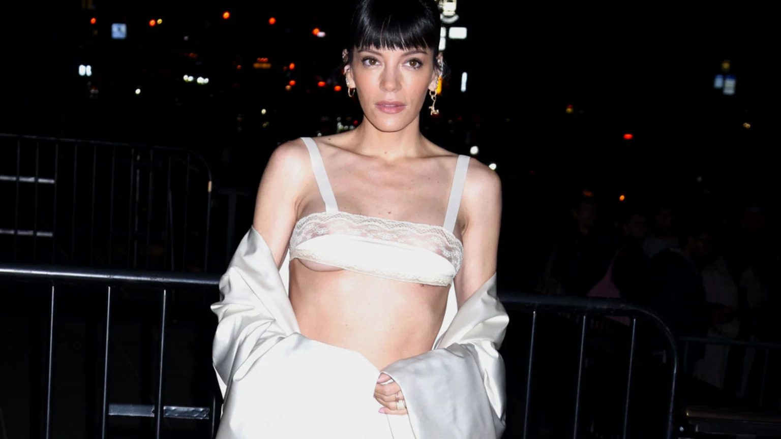 Lily Allen, with dark bangs, looks forward wearing a sheer off-white lace bralette revealing underboob, an open light jacket, and earrings, against a dark, blurry background.