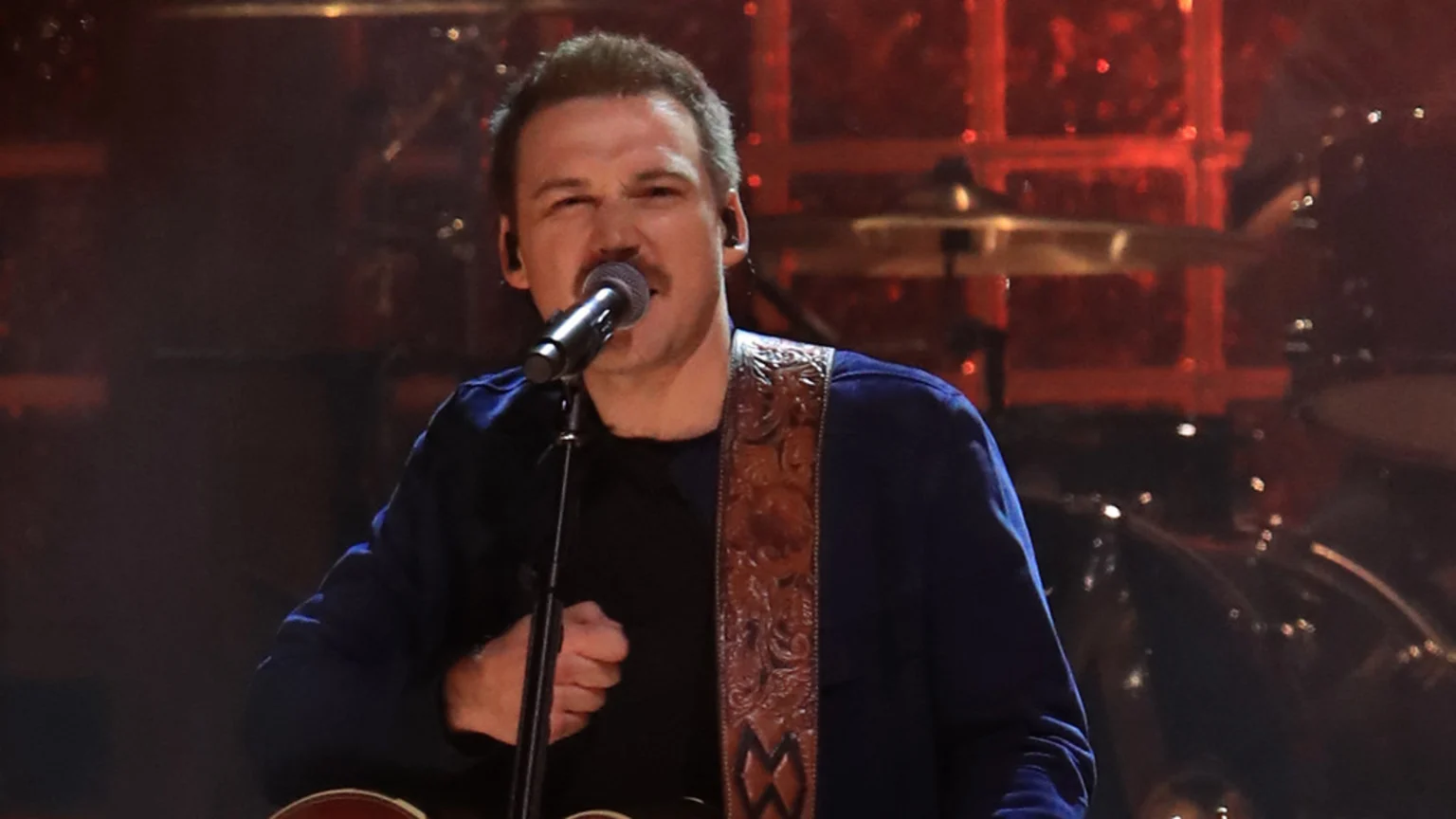 A male singer with a mustache, wearing a blue shirt and a patterned guitar strap, sings into a microphone with a focused expression on a dark stage.