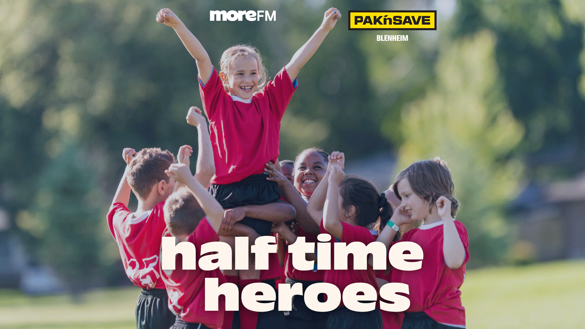 A group of smiling children in soccer uniforms celebrating, with one girl lifted high and her arms raised, with "moreFM", "PAK'nSAVE Blenheim", and "half time heroes" text overlaid.
