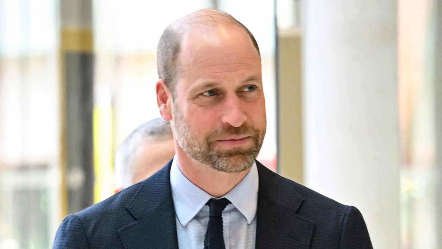 A headshot of Prince William with a beard, wearing a dark blue suit jacket, light blue shirt, and dark tie, looking to his left with a blurred, light background.