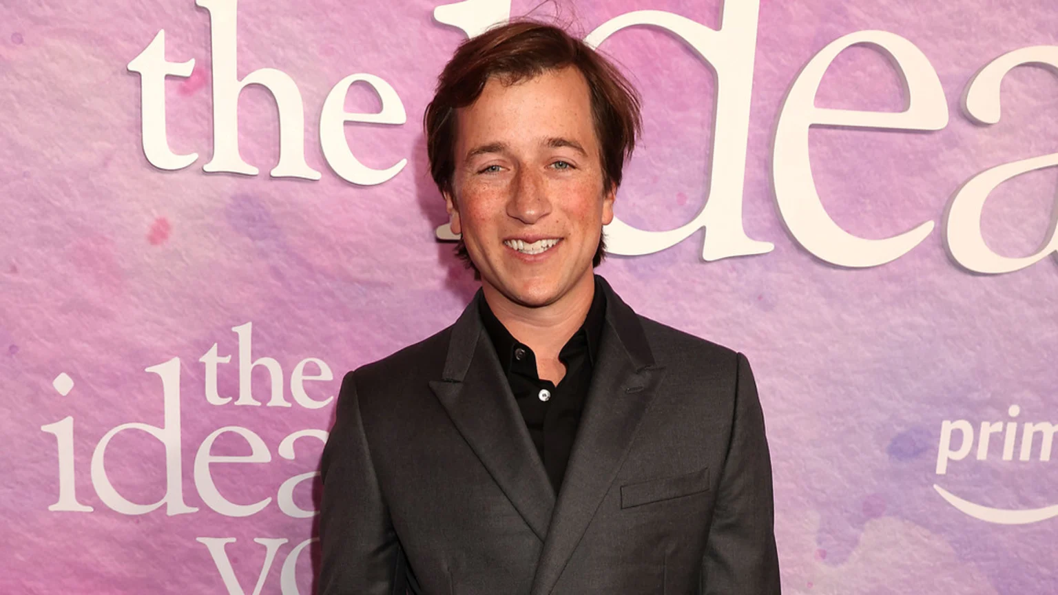 Skyler Gisondo, a young man with light brown hair and freckles, smiles at the camera in a dark gray suit and black shirt against a purple and pink backdrop with white text.