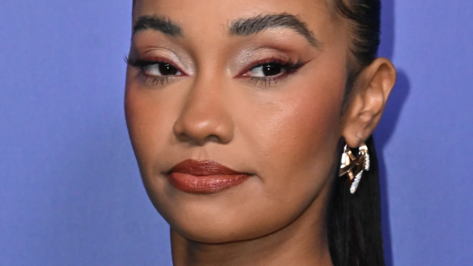 Close-up of a woman with brown skin, dark eyes, and defined eyebrows, wearing reddish-brown winged eyeliner and shimmery lip gloss, with a gold and diamond earring visible against a purple background.