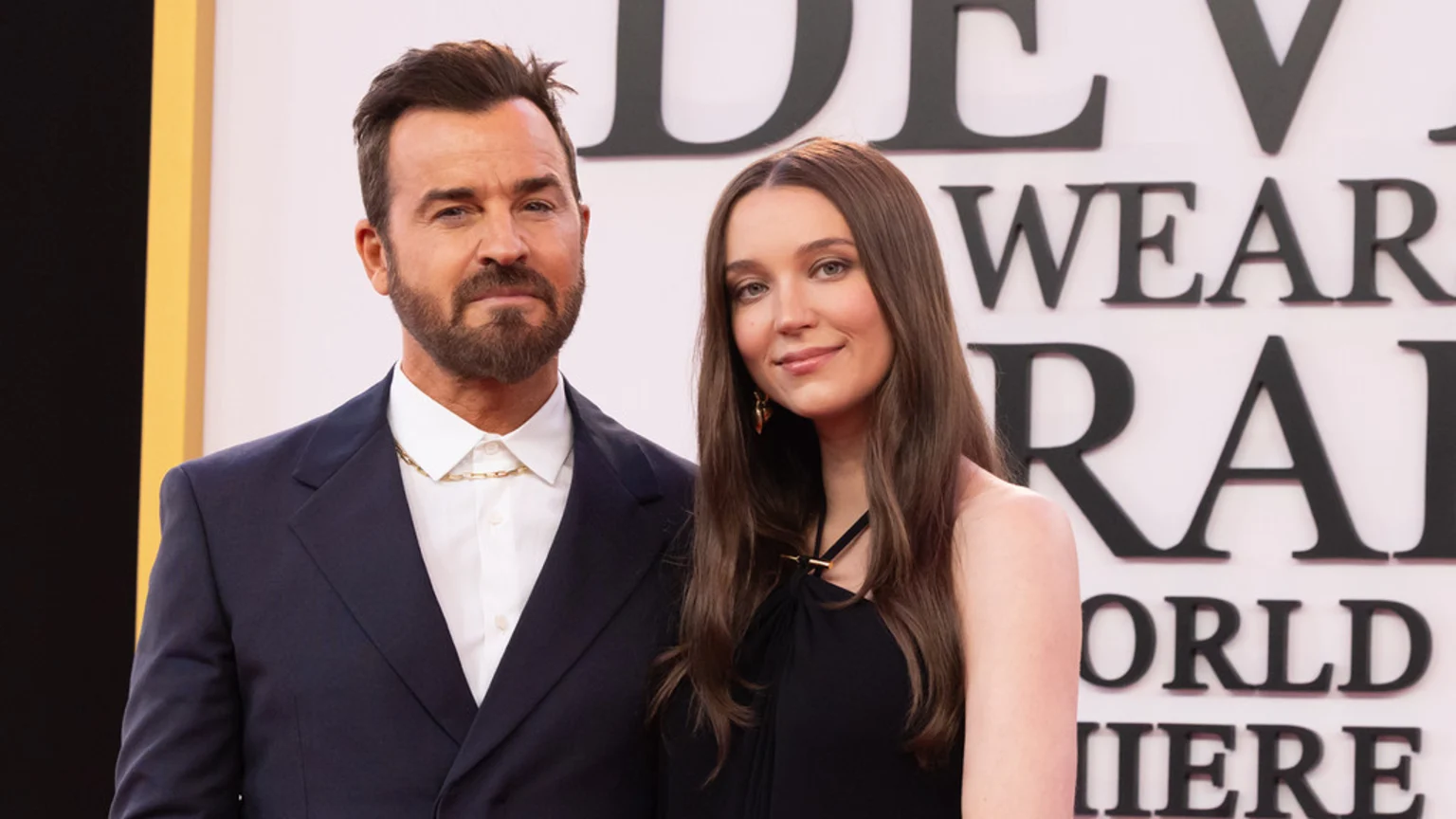 Justin Theroux, in a dark suit and gold chain, stands next to a woman with long brown hair in a black dress, both smiling at an event.