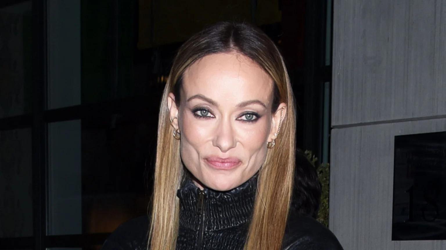A close-up of Olivia Wilde shows her with long blonde hair, dark eye makeup, and a black zippered top, looking forward with distinct lines around her mouth and cheeks.