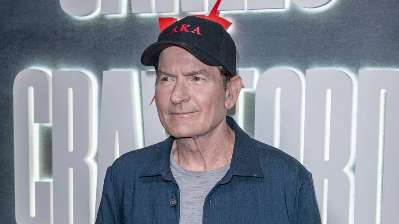 Charlie Sheen in a black cap with red "AKA" text, a blue shirt, and a grey t-shirt, stands in front of large illuminated white letters and a red graphic.
