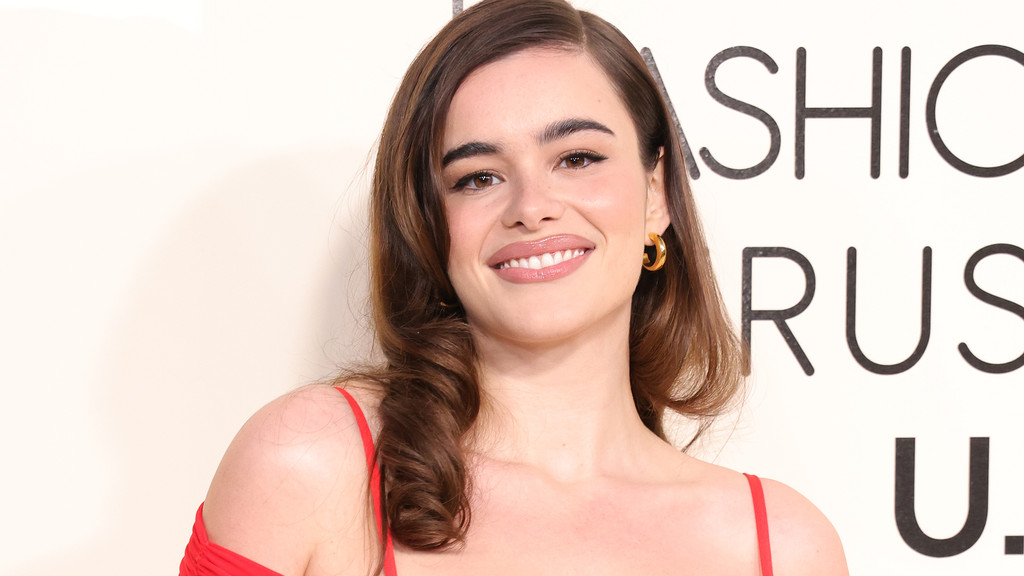 A smiling young woman with brown wavy hair, wearing a red top and gold hoop earrings, looks at the camera against a white background with partial black letters.